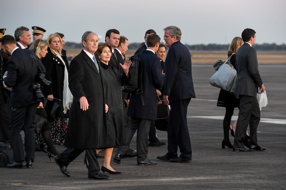 A group of people are all walking to the right. In front are the former president George W. Bush and his wife Laura. They are all dressed in dark suits and winter coats.