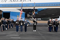 Military members are performing several roles on the flightline in front of Air Force One. In the middle, a color guard is carrying rifles over the shoulders and two members are carrying the US Flag and the flag of the US President