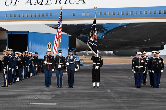 Military members are performing several roles on the flightline in front of Air Force One. In the middle, a color guard is carrying rifles over the shoulders and two members are carrying the US Flag and the flag of the US President