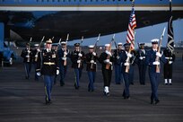 Service members are marching away from Air Force One on a flightline while carrying ceremonial rifles and two members are carrying the US flag and the Flag of the US President.