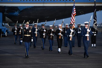 Service members are marching away from Air Force One on a flightline while carrying ceremonial rifles and two members are carrying the US flag and the Flag of the US President.