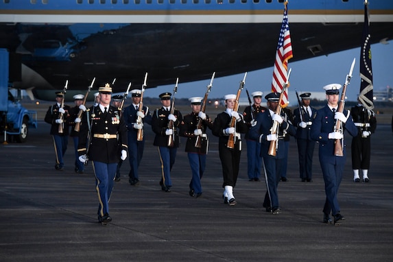 Service members are marching away from Air Force One on a flightline while carrying ceremonial rifles and two members are carrying the US flag and the Flag of the US President.