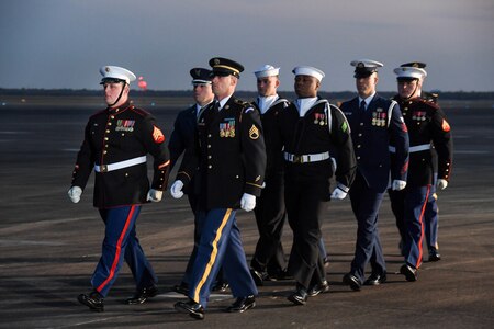 Service members in uniform from each branch of the military are marching down a flightline.