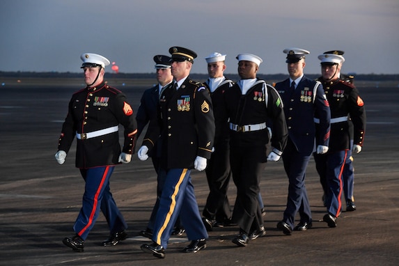 Service members in uniform from each branch of the military are marching down a flightline.
