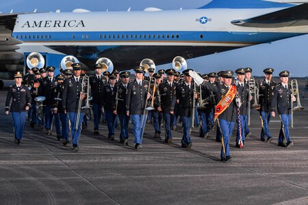 Members of an Army Band are marching near Air Force One on a flightline. The drum major of the band is holding up one hand. The other members are holding various instruments down at their sides.