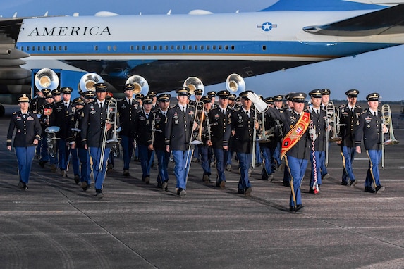 Members of an Army Band are marching near Air Force One on a flightline. The drum major of the band is holding up one hand. The other members are holding various instruments down at their sides.