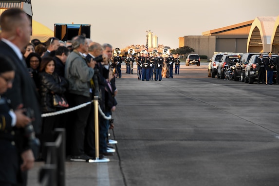 People are standing behind a rope barrier as service members in the distance are preparing to march down a flightline for a funeral procession.