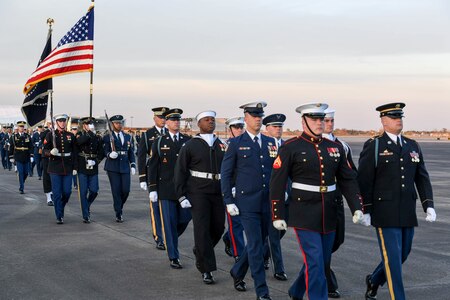 Various service members are marching down a flightline. Some toward the back of the group are carrying ceremonial rifles, as well as the US flag and the flag of the US President.