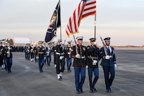 Military members from various branches are marching on a flightline. They are carrying ceremonial rifles as well as the US flag and the flag and of the US President.