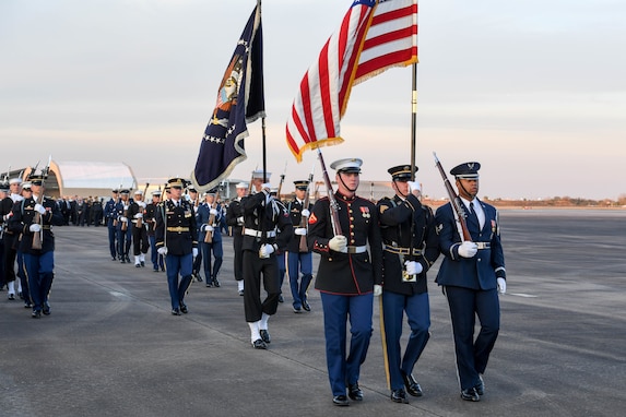 Military members from various branches are marching on a flightline. They are carrying ceremonial rifles as well as the US flag and the flag and of the US President.