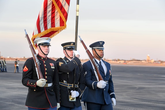 A Marine, an Army Soldier and an Air Force Airman are marching across a flightline carrying rifles and the US flag.