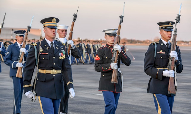 Service members from various branches of the military are marching. The member in front is holding a sword up against his right shoulder, the others are carrying ceremonial rifles in front of them.