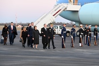Several people in suits and winter coats are being escorted by an Army general officer past a row of service members in uniform holding ceremonial rifles down at their side. In the background is Air Force One.