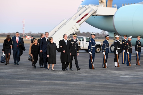 Several people in suits and winter coats are being escorted by an Army general officer past a row of service members in uniform holding ceremonial rifles down at their side. In the background is Air Force One.