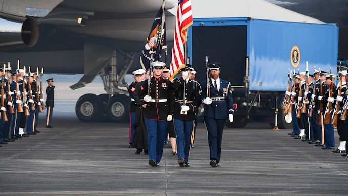 Military members carrying the US flag and flag of the US President are marching away from a light blue truck that is parked next to Air Force Band. They are flanked by other service members who are holding ceremonial rifles in front of them,