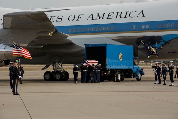 Military members are loading a casket into a light blue colored truck in front of Air Force One. Other military members are holding the US Flag and flag of the US President, as well as ceremonial rifles.