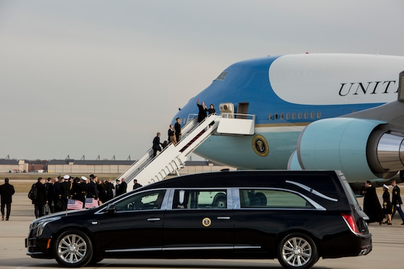 A hearse with the seal of the President of the US on the door and US flag affixed to the hood is parked in front of Air Force One (large plane with light blue and white) as several people board the plane using steps up to the door.