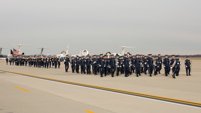 A long processional of people in military uniforms are marching down a flightline. In the front are Air Force Band members in dark blue uniforms carrying their instruments. Behind them are members of the honor guard, and the a color guard carrying the US and US President's flag are at the rear of the group.