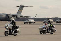 Two police on motorcycles are riding down a flightline. In the distance is a large grey cargo plan, and several vehicles, including a hearse.