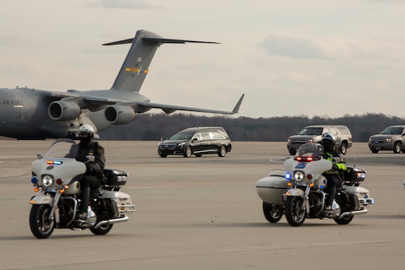 Two police on motorcycles are riding down a flightline. In the distance is a large grey cargo plan, and several vehicles, including a hearse.