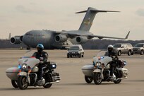 Two police officers on motocycles are riding across a flightline with a large grey cargo plane in the distance and several other vehicles, including a hearse, following them.