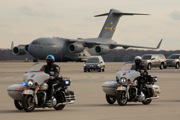 Two police officers on motocycles are riding across a flightline with a large grey cargo plane in the distance and several other vehicles, including a hearse, following them.