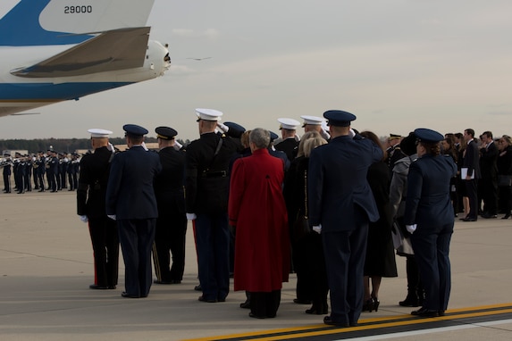 People are turned away from the camera, looking toward a plane on the flightline. Some are in uniform and are saluting. The tail of the plane is white and light blue. It is Air Force One. It the far distance, a band in uniform is visible.