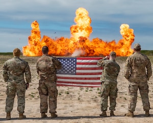 Four Airmen stand in a field, two airmen in the center hold a flag, while they watch an explosion.