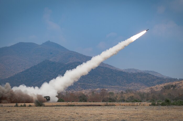 A vehicle-fired missile leaves a trail of smoke as it launches over a training range during daylight.