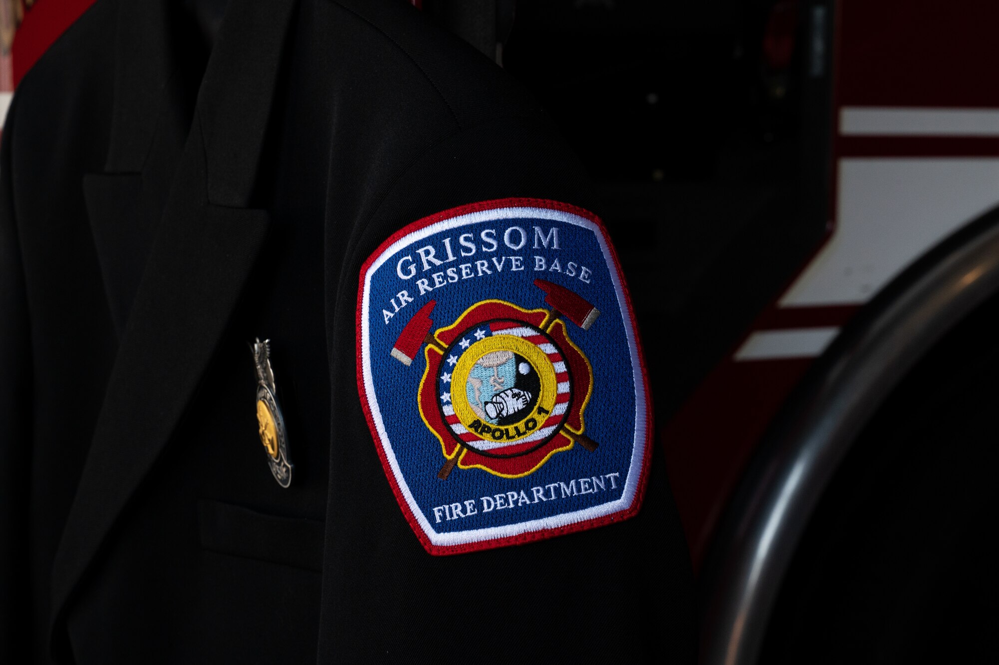 A patch that reads "GRISSOM AIR RESERVE BASE FIRE DEPARTMENT" with an emblem that displays the Apollo 1 with an American flag visible in the background, accompanied by fire axes on either side.