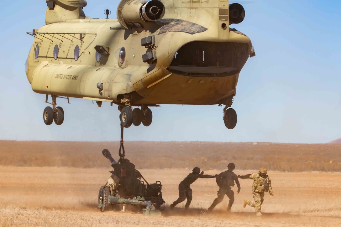 A large helicopter uses a cable to lift a heavy weapon in a field as three soldiers prepare to get out of the way.