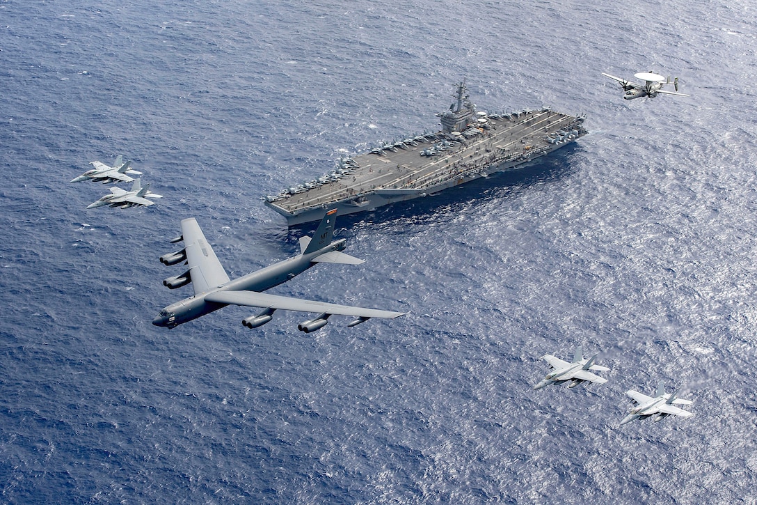 Six aircraft fly above an aircraft carrier at sea.