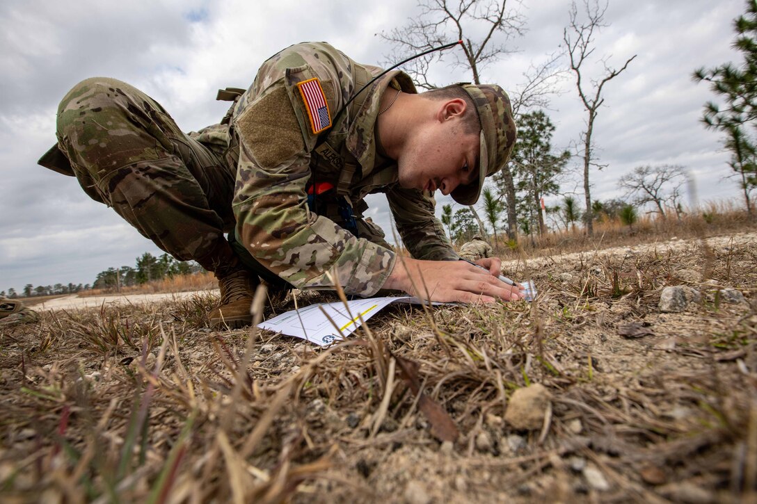 A soldier crouches in a field and examines a piece of paper.