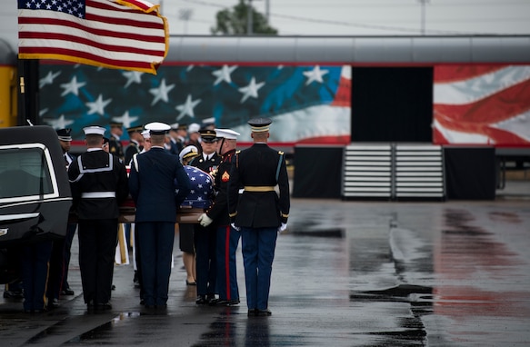 Service members from each branch of the military dressed in their ceremonial uniforms are lifting a flag-draped coffin out of a hearse. The US Flag is waving on the left side of the picture. There is a train car that has the US flag (red white and blue) on it in the background. The ground is wet asphalt.