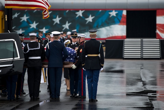 Service members from each branch of the military dressed in their ceremonial uniforms are lifting a flag-draped coffin out of a hearse. There is a train car that has the US flag (red, white and blue) on it in the background. The ground is wet asphalt.