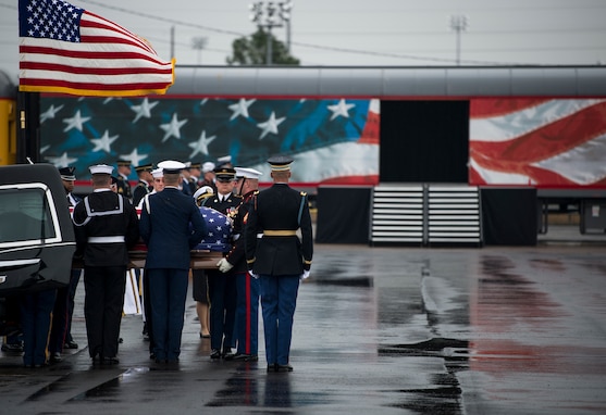 Service members from each branch of the military dressed in their ceremonial uniforms are lifting a flag-draped coffin out of a hearse. There is a train car that has the US flag (red white and blue) on it in the background. The ground is wet asphalt.