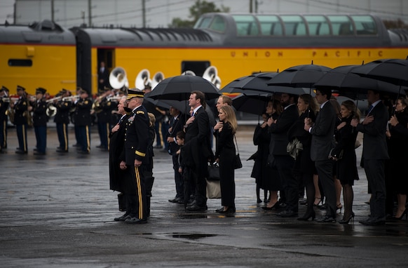 People dressed in dark clothing are standing with umbrellas on a wet asphalt pavement while looking toward the left side of the picture. There is a military band dressed in dark uniforms performing and a yellow train in the background.