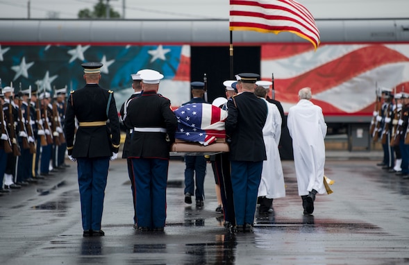 Service members from each branch are dressed in ceremonial uniforms standing in two long parallel rows. In the background is a train with red, white and blue colors, and a flag-drapped coffin is being carried onto the train car.