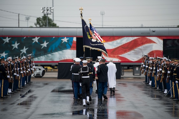 Service members from each branch are dressed in ceremonial uniforms standing in two long parallel rows. In the background is a train with red, white and blue colors, and a flag-drapped coffin is being carried onto the train car.