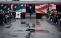 Service members from each branch are dressed in ceremonial uniforms standing in two long parallel rows. In the background is a train with red, white and blue colors, and a flag-drapped coffin is being carried onto the train car.