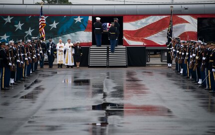 Service members from each branch are dressed in ceremonial uniforms standing in two long parallel rows. In the background is a train with red, white and blue colors, and a flag-drapped coffin is being carried onto the train car.