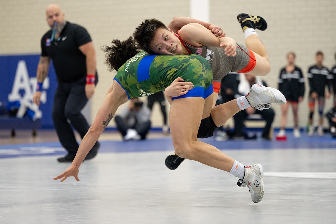 A wrestler performs a double leg takedown move on another wrestler during competition as a referee watches.