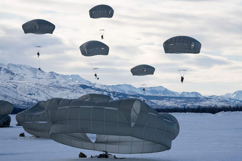 Paratroopers prepare to land on snowy ground after jumping from a military aircraft on a gray, cloudy day.