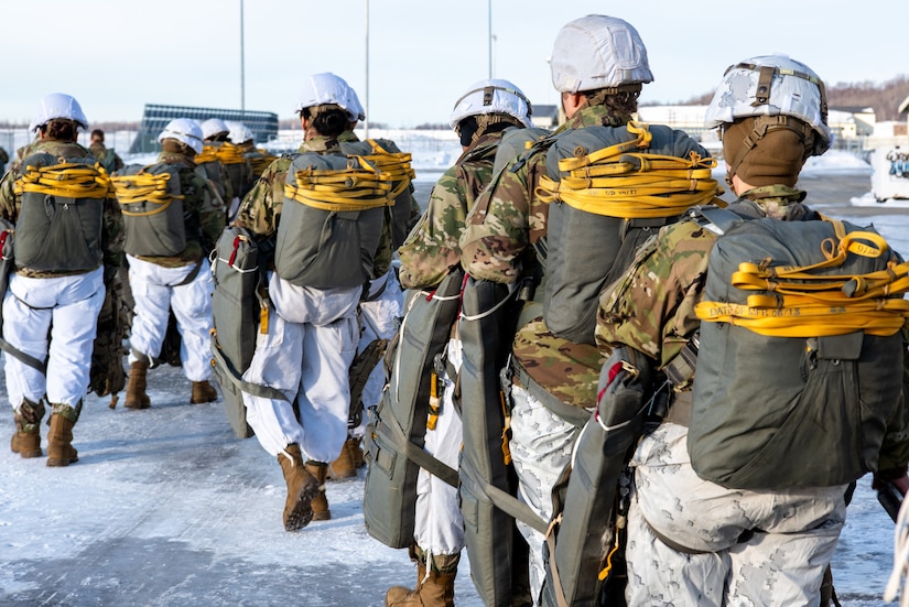 Army paratroopers stand in a line as they prepare to board a military aircraft during daylight.