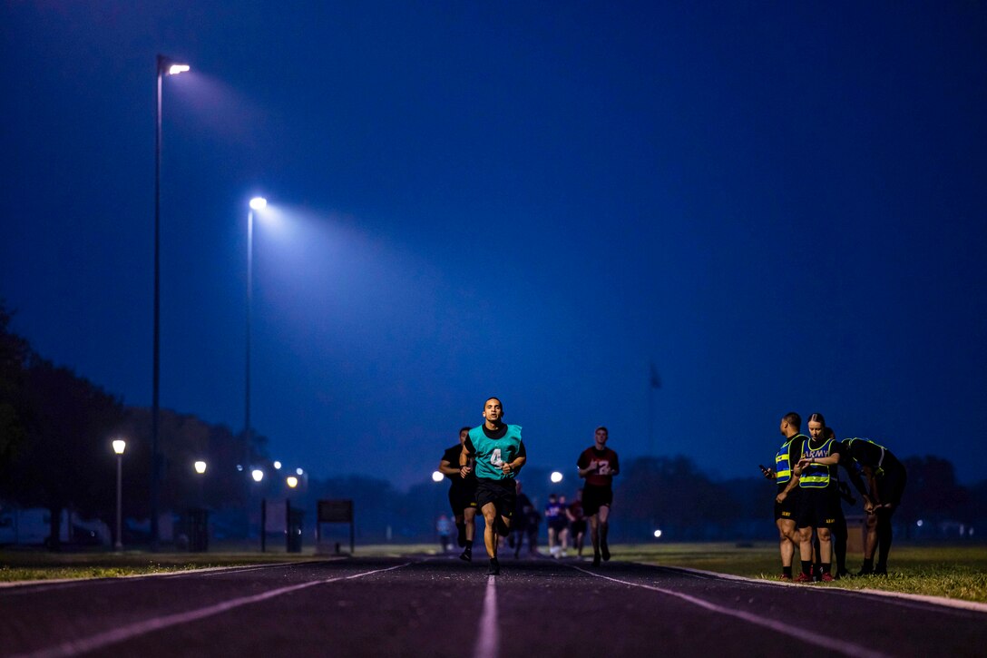 Soldiers run through fog on a track under spotlights as fellow soldiers stand on the sideline preparing to run.