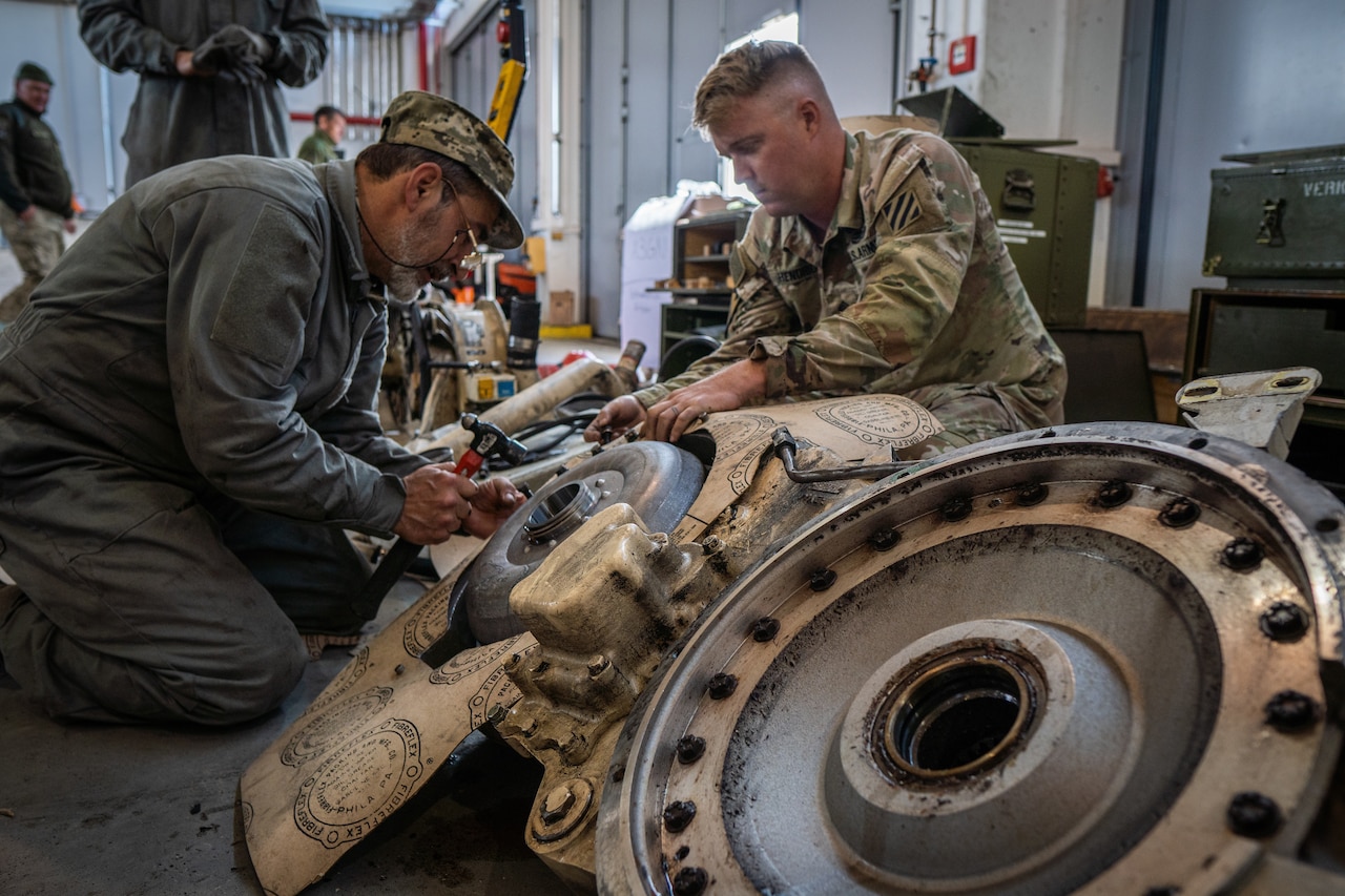 A uniformed service member and a man work on a piece of military equipment.