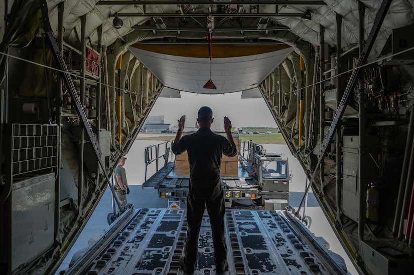 A man, standing in the bay of an airplane signals for another truck to deliver a wooden box.