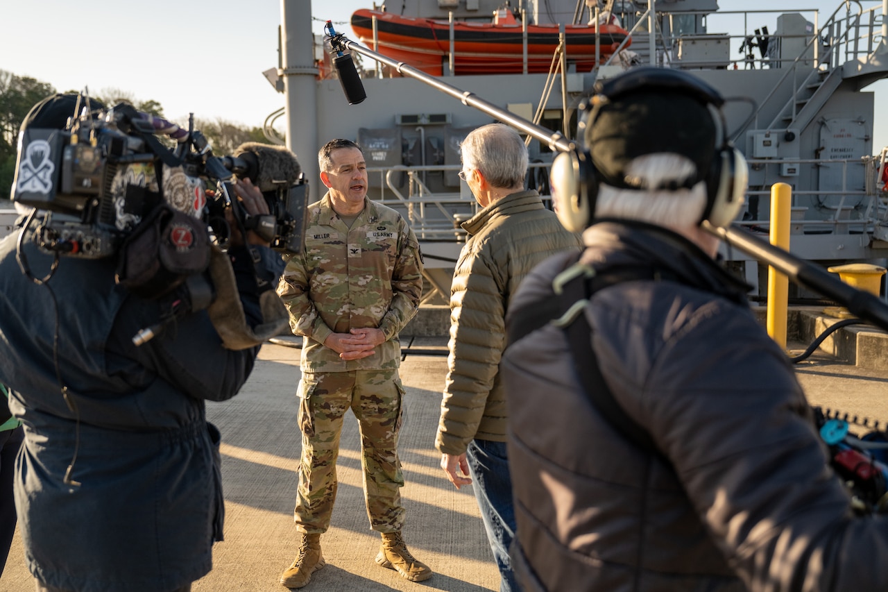 A soldier in uniform speaks to a camera crew on a pier.