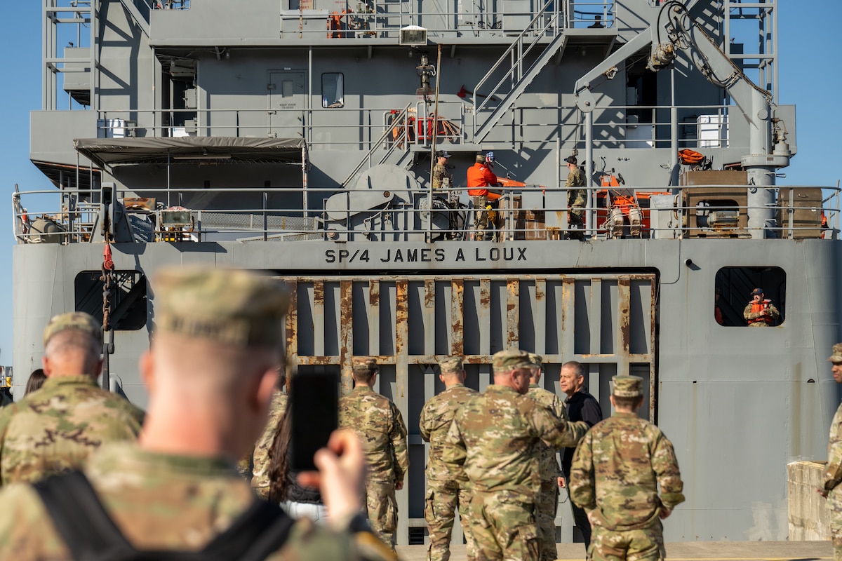 Soldiers in uniform stand on a pier as crews work aboard a military vessel.