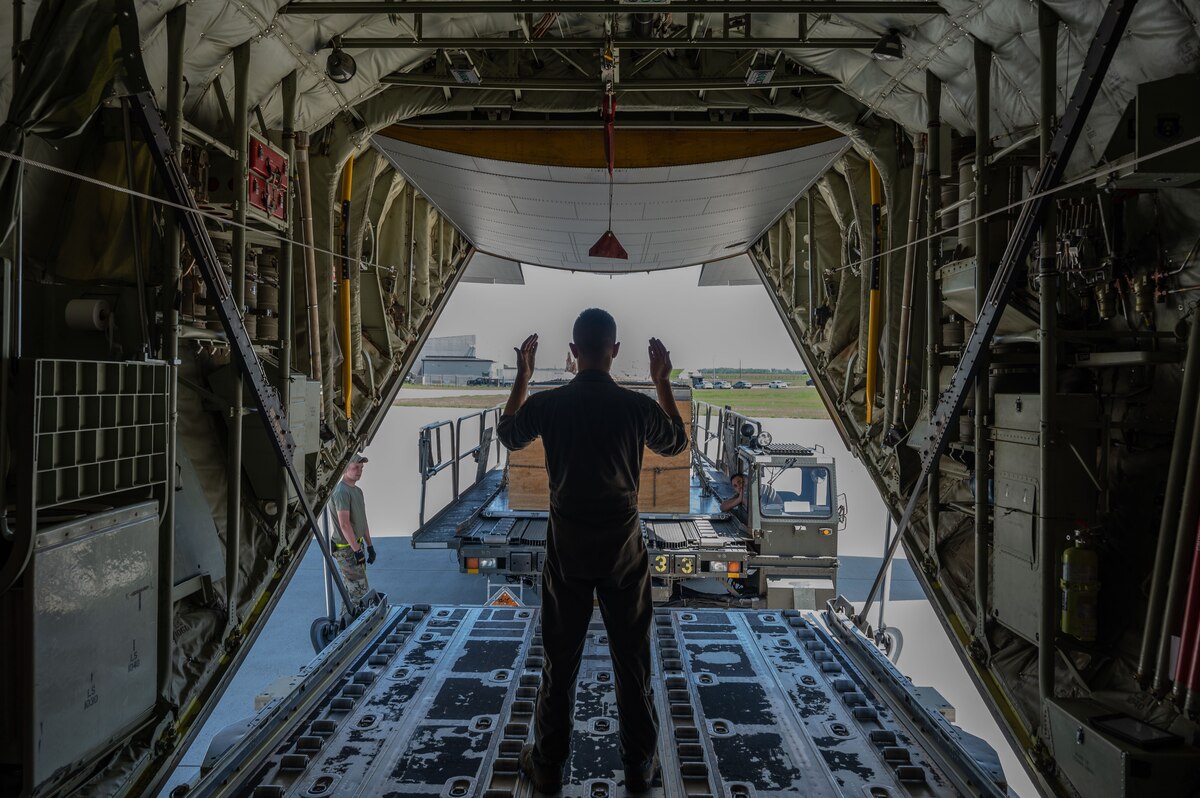 A man, standing in the bay of an airplane signals for another truck to deliver a wooden box.